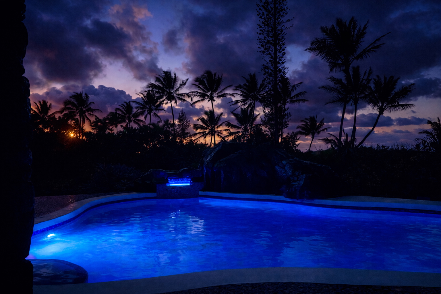 Oceanfront pool glowing beneath a deep sunset and palm silhouettes