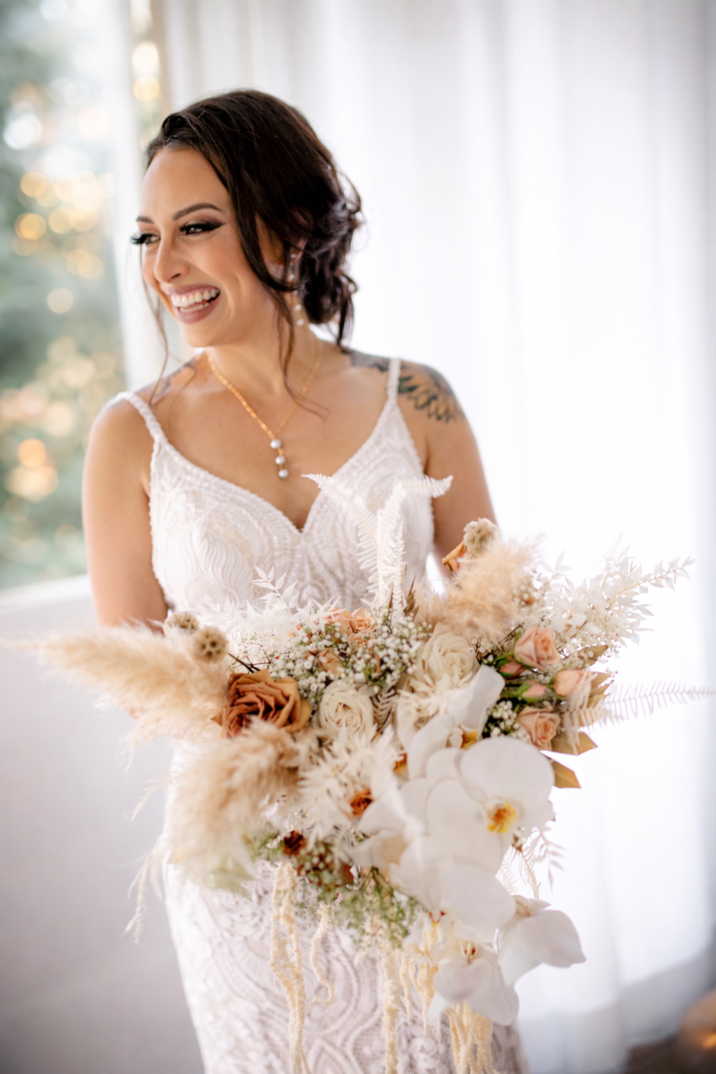 Bride laughing in a lace gown while holding a large pampas and orchid bouquet in soft window light